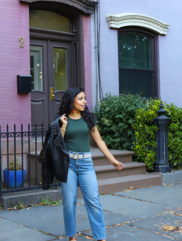 latinx woman wearing denim jeans, a green top, leather jacket in front of a bright pink building